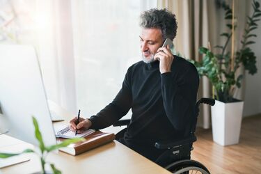 Mann in Rollstuhl am Schreibtisch telefoniert vor Bildschirm