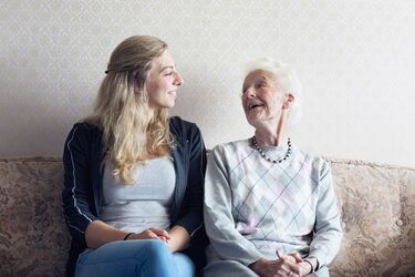junge Frau sitzt zuusammen mit älterer Dame auf Couch und lacht herzlich